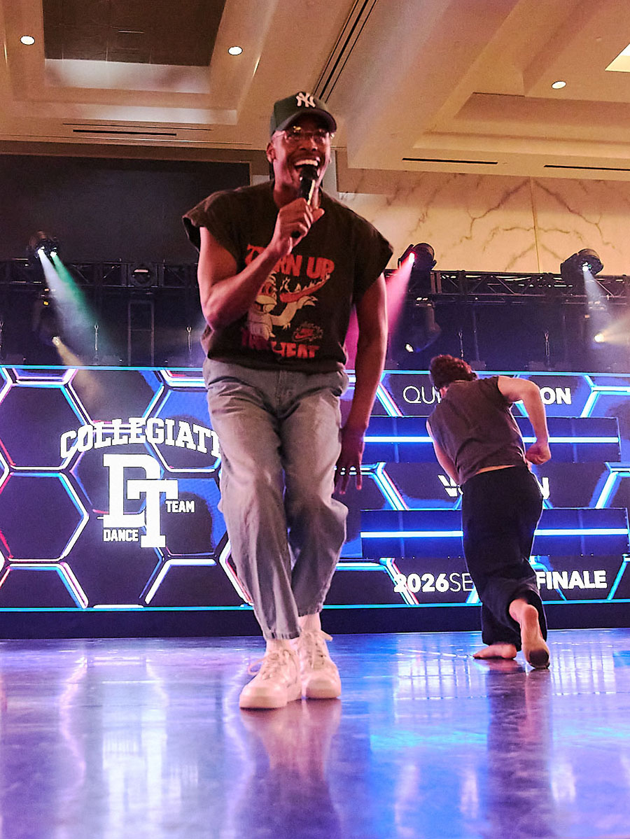 A performer with a microphone dancing in front of the Collegiate Dance Team Night stage backdrop